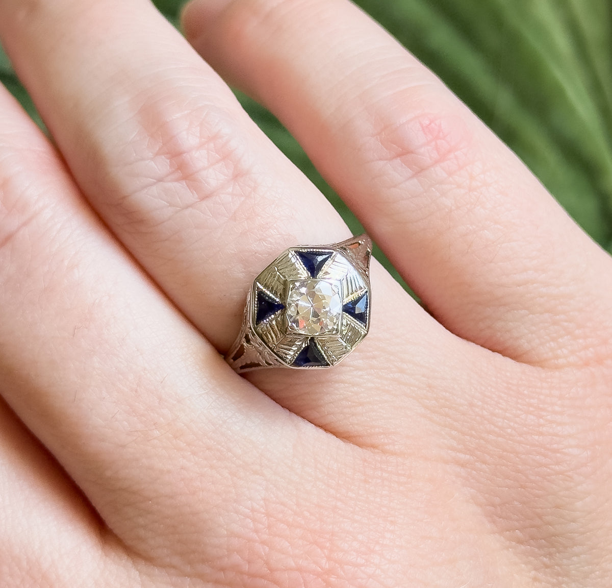Close overhead view on the hand, the ring filling much of the finger's width so the central diamond's facets and the four triangular blue stones show their scale.