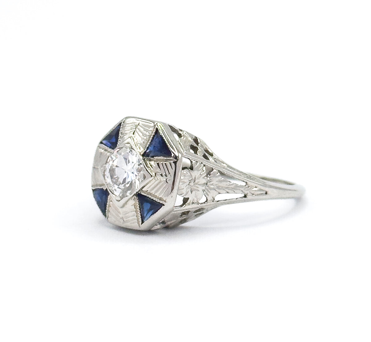 Angled three-quarter view on white, the octagonal face turned so the Old European Cut diamond and triangular blue stones are seen alongside the etched floral motif on the shoulder.