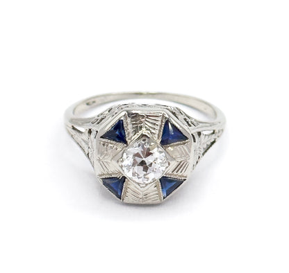 Straight-on overhead view on white, the octagonal face square to the camera so the Old European Cut diamond sits centred within four triangular blue stones and etched rays.