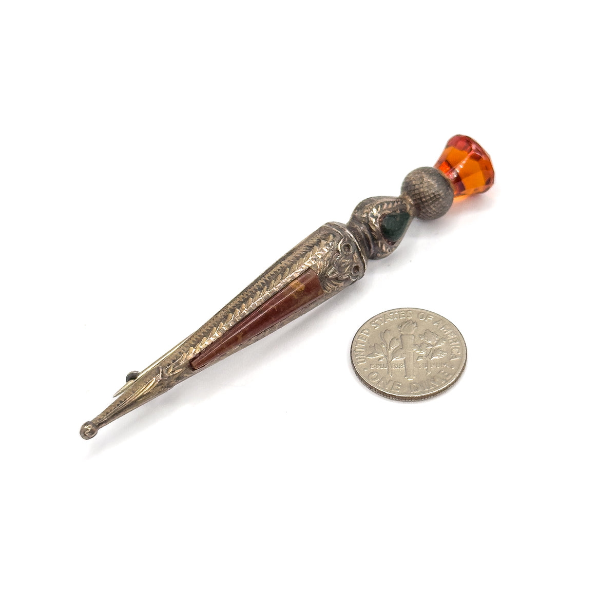 Shown flat on white beside a dime for scale, the thistle head with its faceted orange glass at upper right and the tapering agate-inlaid body running down to the pin point.