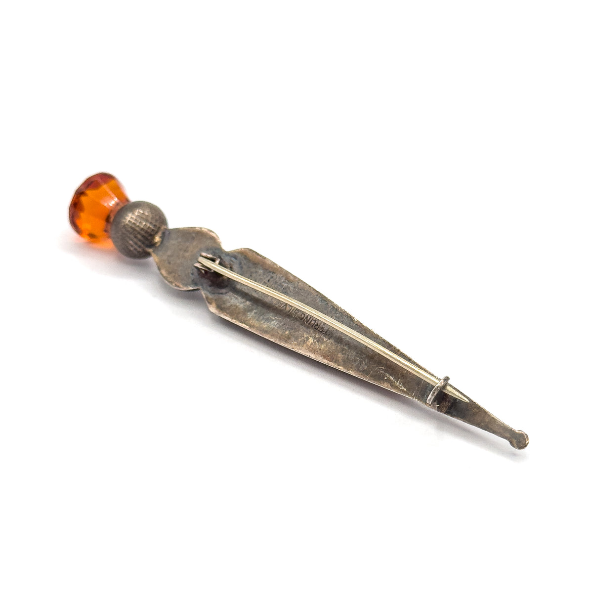 Angled view of the back, the long pin stem crossing the patinated body and hooking into the catch at the tapered tip, with the faceted orange glass showing at the far end.