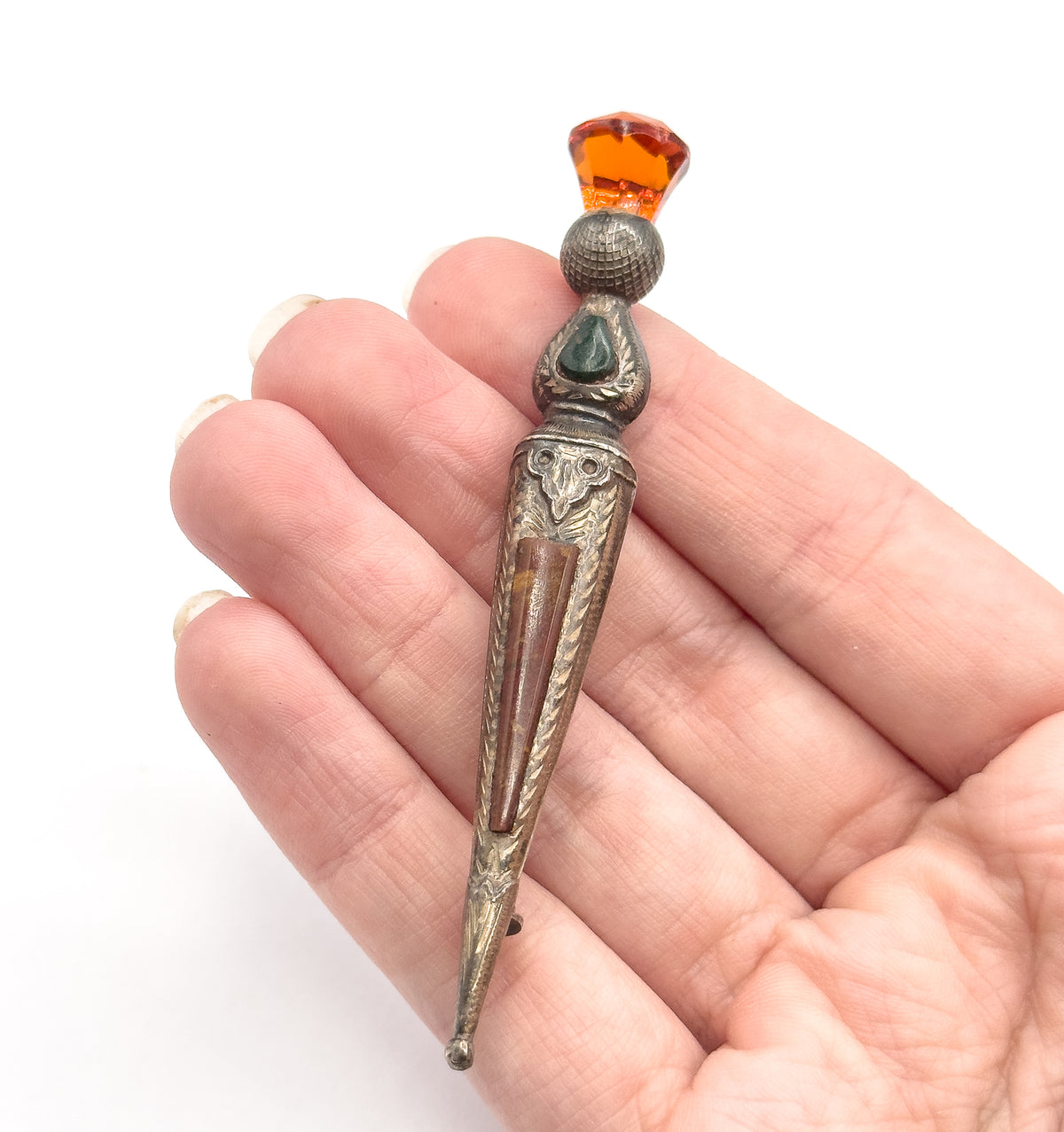 Victorian thistle kilt pin brooch in sterling silver with faceted orange glass and agate inlay, shown resting across an open palm, its tapering pin length spanning the fingers to give scale.