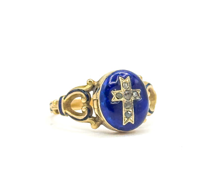 Three-quarter view with the locket closed, the deep blue enamel oval facing the camera and its cross of six small faceted stones upright between the scrolled, enamel-accented shoulders.