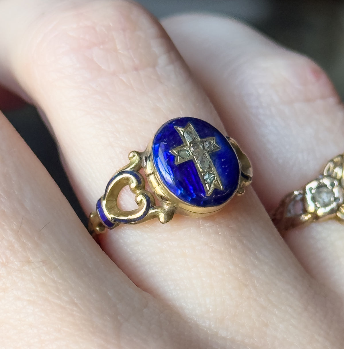 Victorian blue enamel locket ring in 15K gold, its oval face set with a cross of six rose cut diamonds, shown on a finger with openwork enamelled shoulders beside another ring.