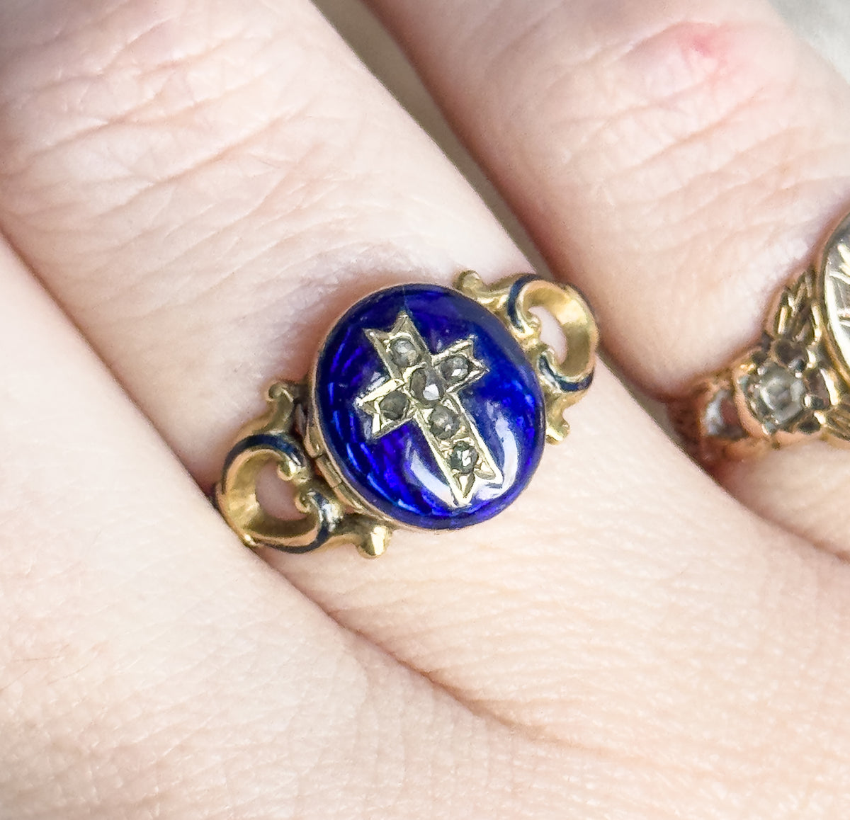 Tight overhead close-up looking straight down at the blue enamel face, where the diamond cross and scrolled gold shoulders sit against the finger's creases for scale.