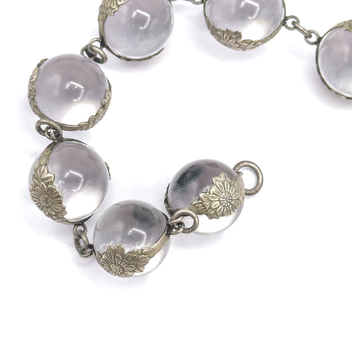 A curving run of rock crystal orbs on white with an open silver clasp ring standing clear at the end of the strand, the floral mountings and small jump rings seen from above.