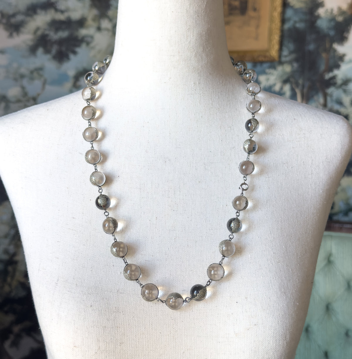 Art Deco pools of light rock crystal necklace in sterling silver, shown at the neck on a cream dress form, its row of round clear orbs held in floral silver mountings with a small clasp at one side.