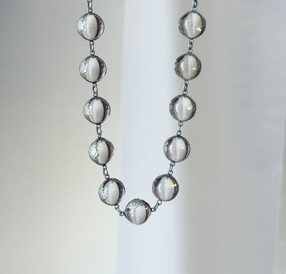 The lower U of the strand hanging free against a pale draped backdrop, each clear orb linked to the next by short silver links and gripped in its floral mounting.