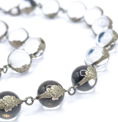 Curving line of orbs on a white background, the nearest spheres reflecting dark in their floral silver mountings while the links and jump rings between them show clearly up close.