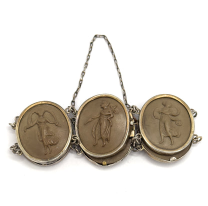 Three carved khaki-brown cameos of draped figures seen from slightly above, their bezels tipped forward so the prong-backed sides show, with the safety chain arching high overhead.