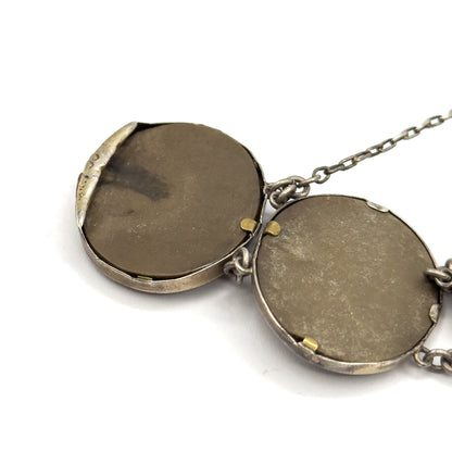Reverse view of two oval settings, showing the flat khaki-brown backs of the cameos held by small prong tabs inside their bezels, with the chain links and safety chain alongside.