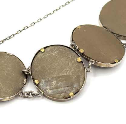 Angled close-up of the reverse, where four flat khaki-brown cameo backs sit in their bezels on small gold-tone prong tabs, with the fine safety chain crossing the plain background above.