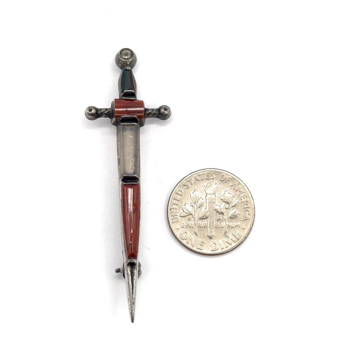 Laid flat beside a dime for scale on white, the sword brooch little longer than two coins, its reddish-brown, grey and dark green cabochons set along blade, crossguard and pommel.