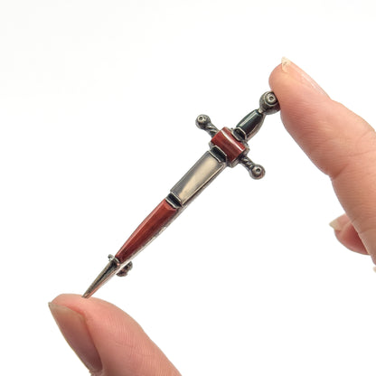 A vintage sterling silver sword brooch set with chalcedony, quartz and bloodstone cabochons, held upright between two fingertips which show its small size.