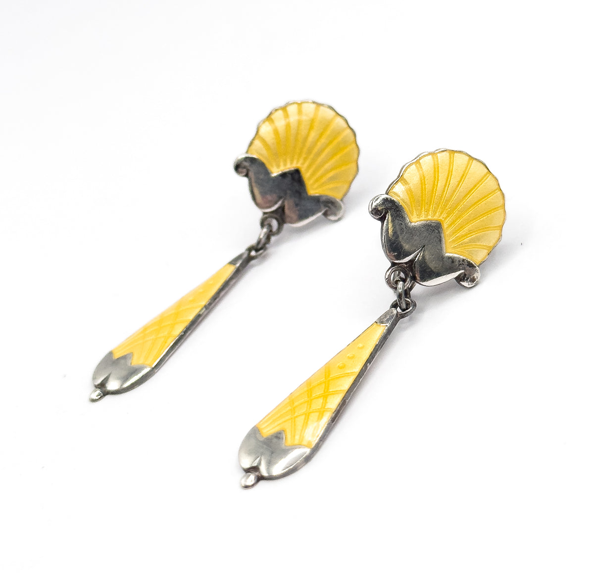 Both earrings lie diagonally on white in a raking three-quarter view, light catching the scrolled silver brackets and jump rings that link each fluted yellow shell to its long tapered drop.