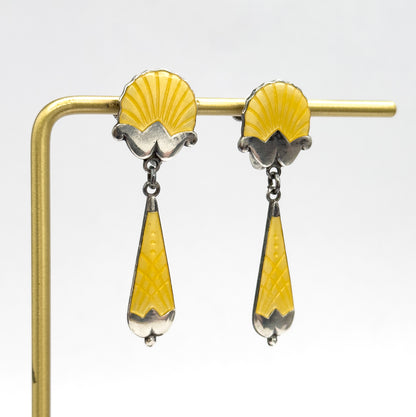Both earrings hang side by side from a slim light-coloured stand, showing the shell tops, small links and long tapered yellow enamel drops in profile against a plain background.