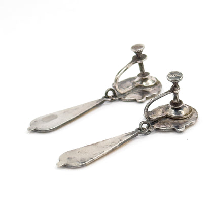 The backs of both earrings face up, showing the screw fittings with threaded posts and knurled discs, one stamped, the shell reverses carrying patina and the plain undersides of the drops.
