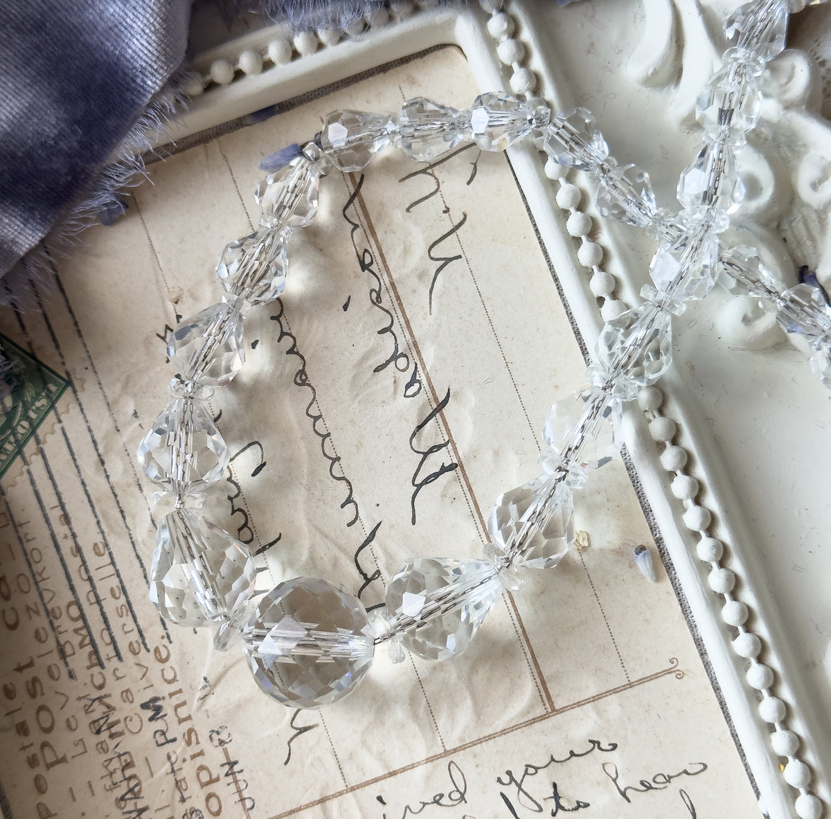 An Art Deco faceted crystal glass bead necklace shown coiled on a handwritten postcard, its graduated colourless beads in assorted shapes catching the light.