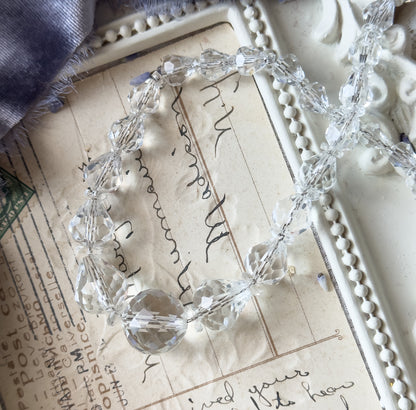 An Art Deco faceted crystal glass bead necklace shown coiled on a handwritten postcard, its graduated colourless beads in assorted shapes catching the light.