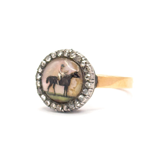 Front view tilted slightly left on white, the painted horse and rider under the domed crystal catching pink and green tones, ringed by the silver-set rose cut diamond halo above the gold shank.