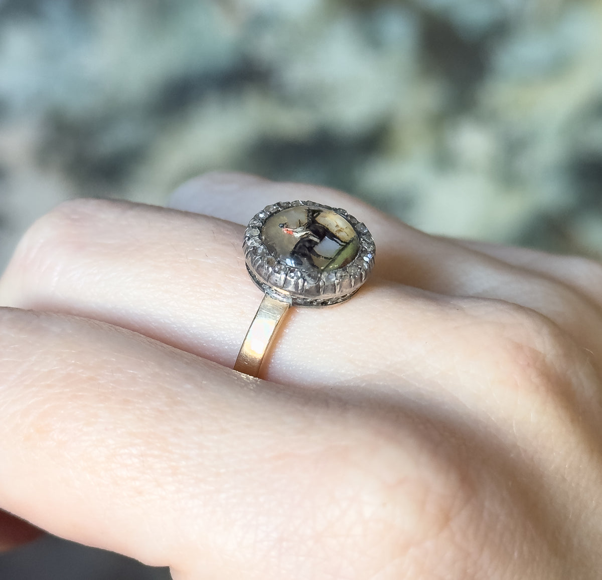 Angled side view on the finger, where the slim yellow gold shank rises to meet the deep silver-topped bezel and its ring of small diamonds, against a soft green blurred background.