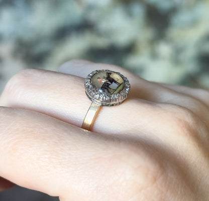 Angled side view on the finger, where the slim yellow gold shank rises to meet the deep silver-topped bezel and its ring of small diamonds, against a soft green blurred background.