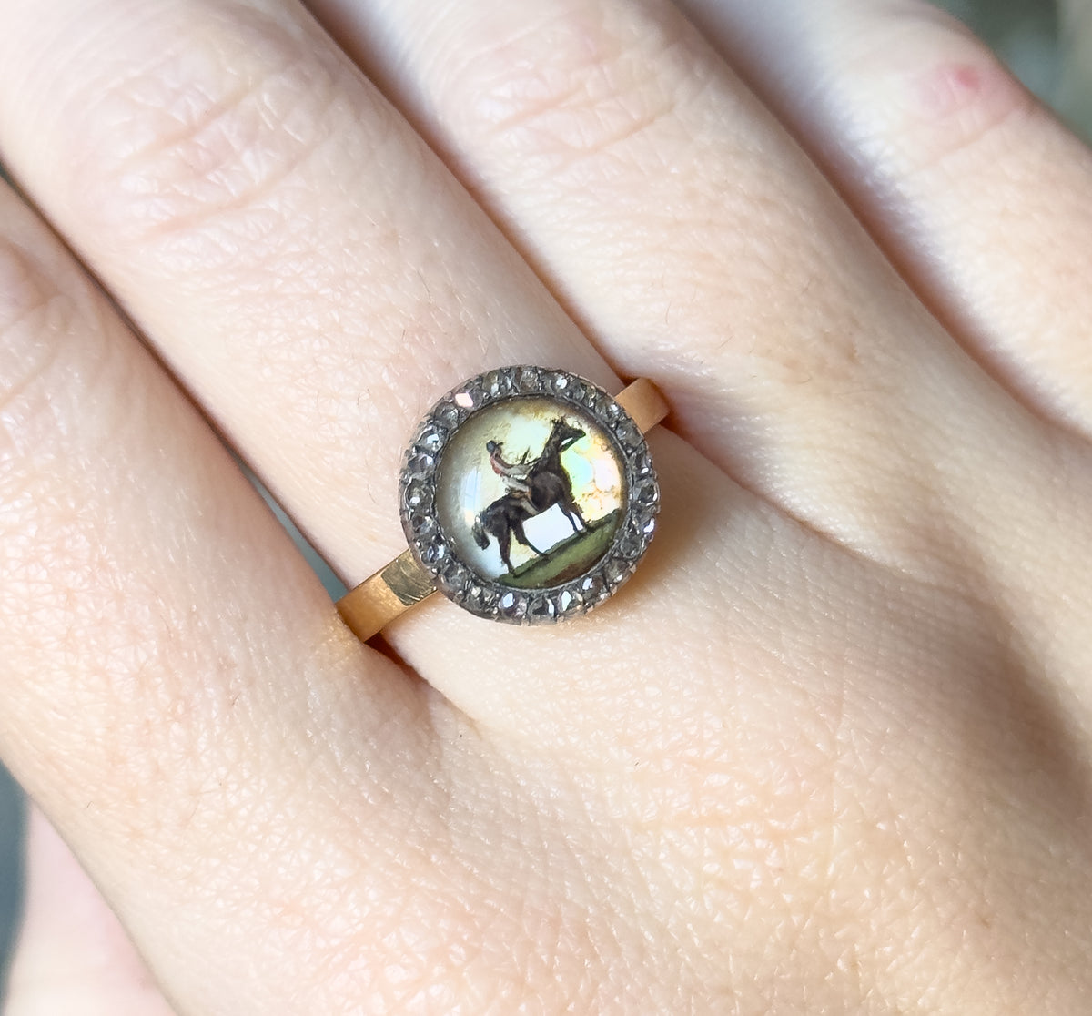 Shown on a finger tilted toward the light, so the abalone backing flashes pale green and pink around the horse and rider while the flat gold shank crosses the finger below the bezel.