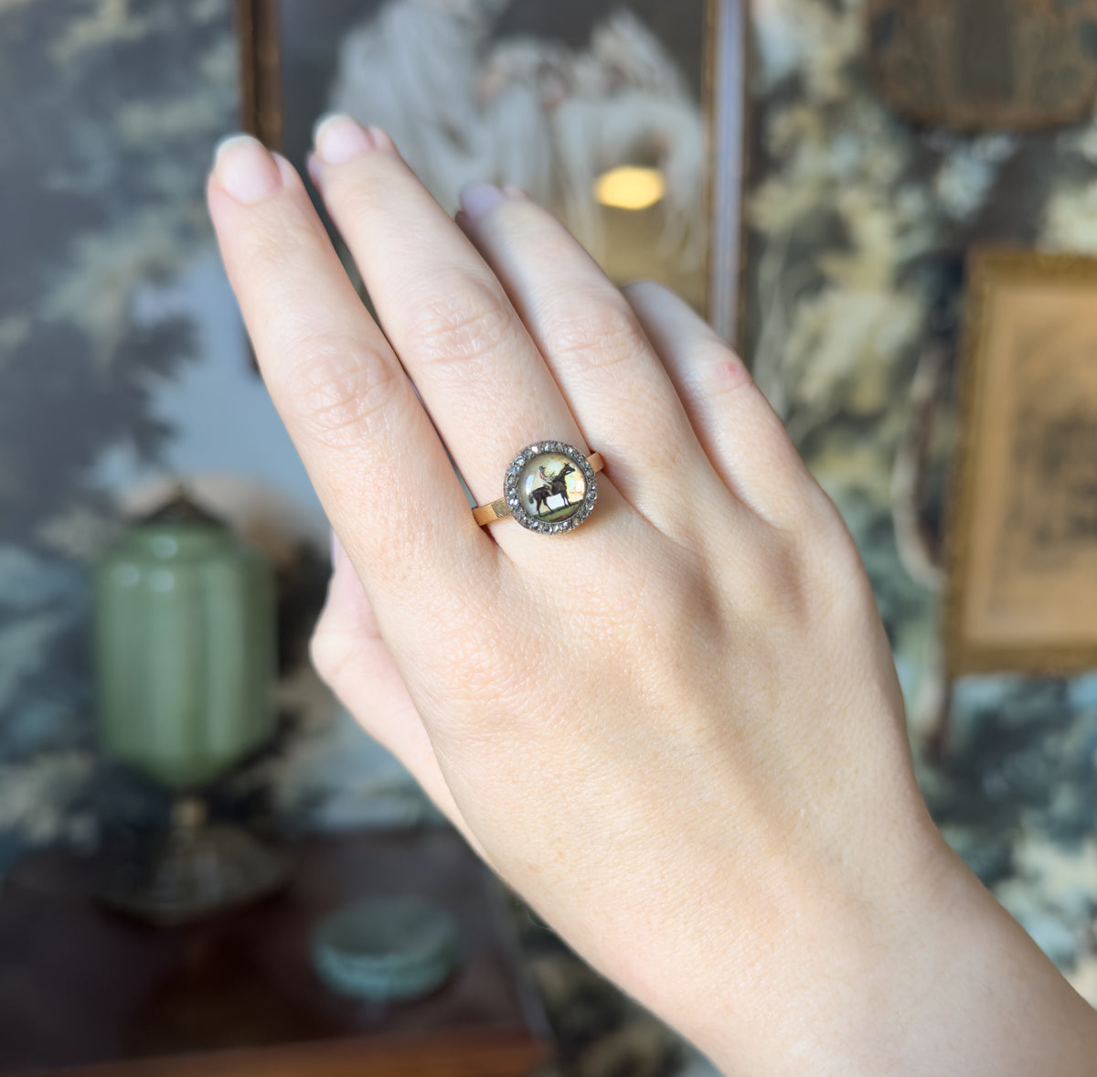 Full view of the hand raised against a blurred room interior, the ring sitting on the ring finger so its round bezel and halo read small against the knuckle, with the flat gold shank below.