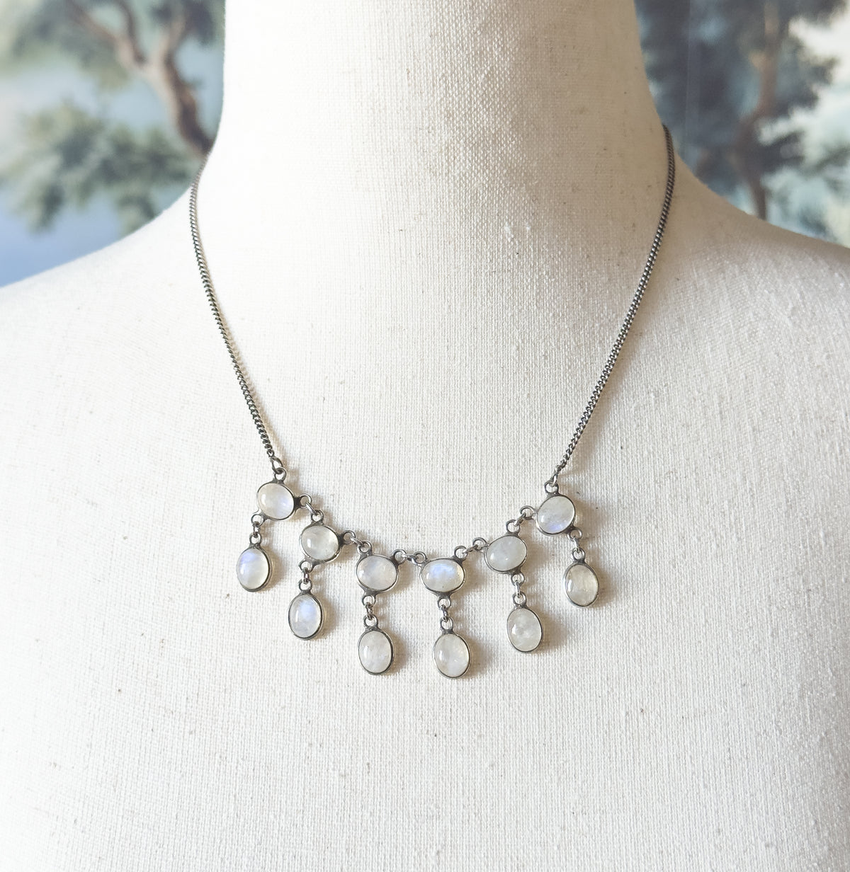 Closer from above on the linen bust, the fine curb-link chain traced to where each upper bezel meets a longer oval drop, the moonstones catching cool blue-green glints.