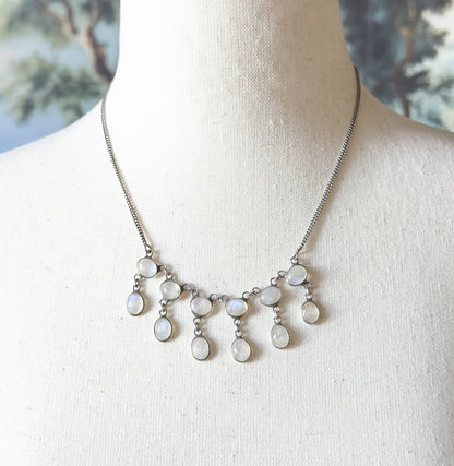 Closer from above on the linen bust, the fine curb-link chain traced to where each upper bezel meets a longer oval drop, the moonstones catching cool blue-green glints.