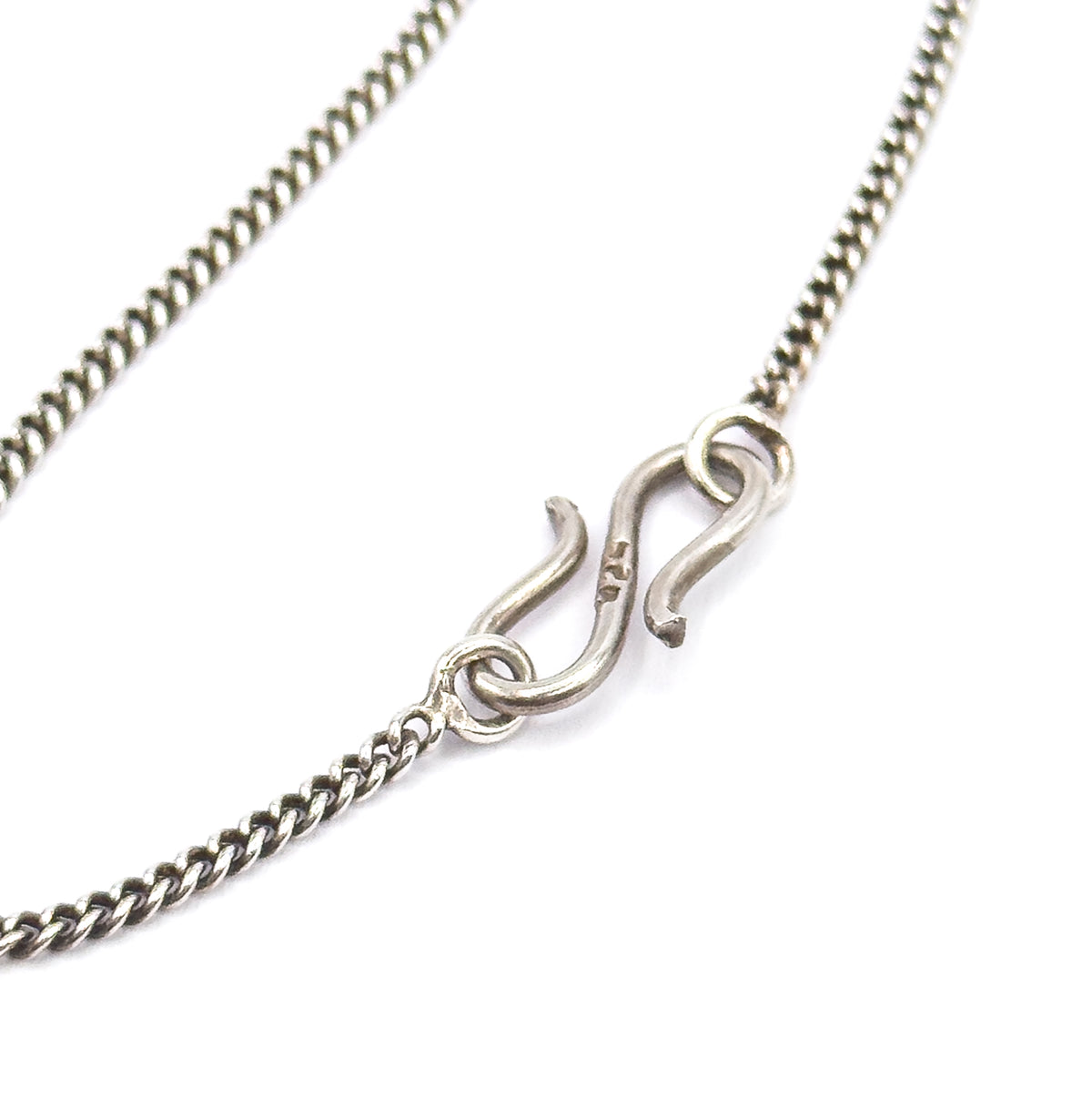 Macro on white of the S-shaped hook clasp and its jump ring linking the two ends of the fine curb-link chain, the darkened patina settled between the links.