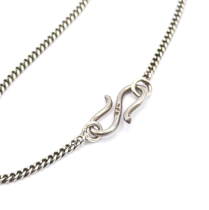 Macro on white of the S-shaped hook clasp and its jump ring linking the two ends of the fine curb-link chain, the darkened patina settled between the links.