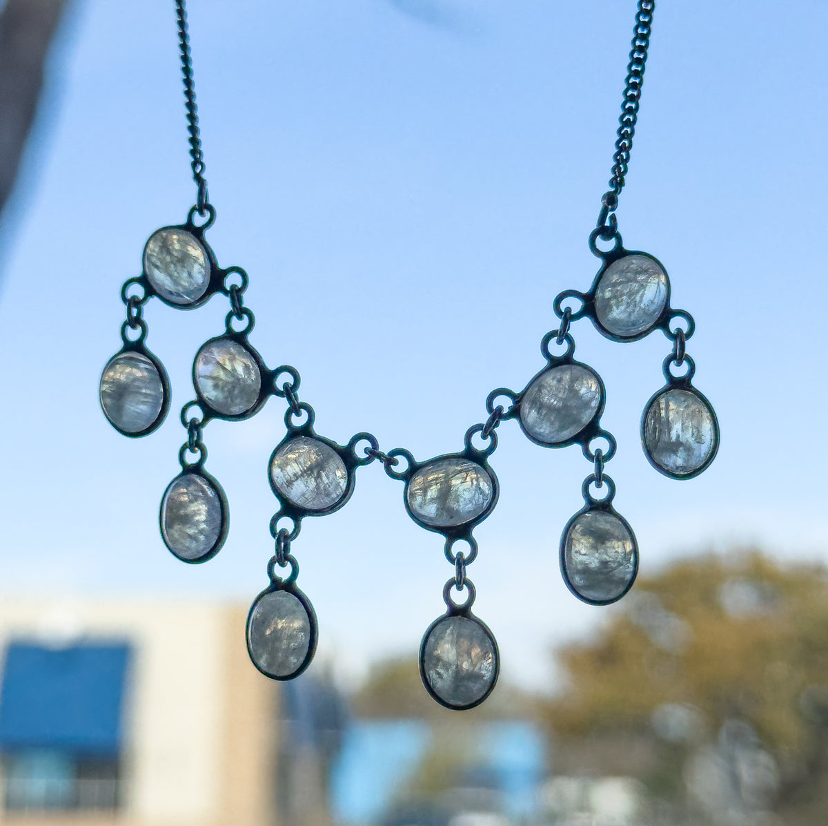 Edwardian moonstone fringe necklace in sterling silver, held up against a bright sky so the bezel-set oval cabochons and their drops hang free, the darkened patina outlining each stone.