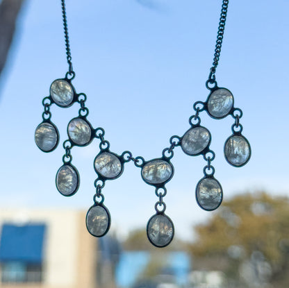 Edwardian moonstone fringe necklace in sterling silver, held up against a bright sky so the bezel-set oval cabochons and their drops hang free, the darkened patina outlining each stone.