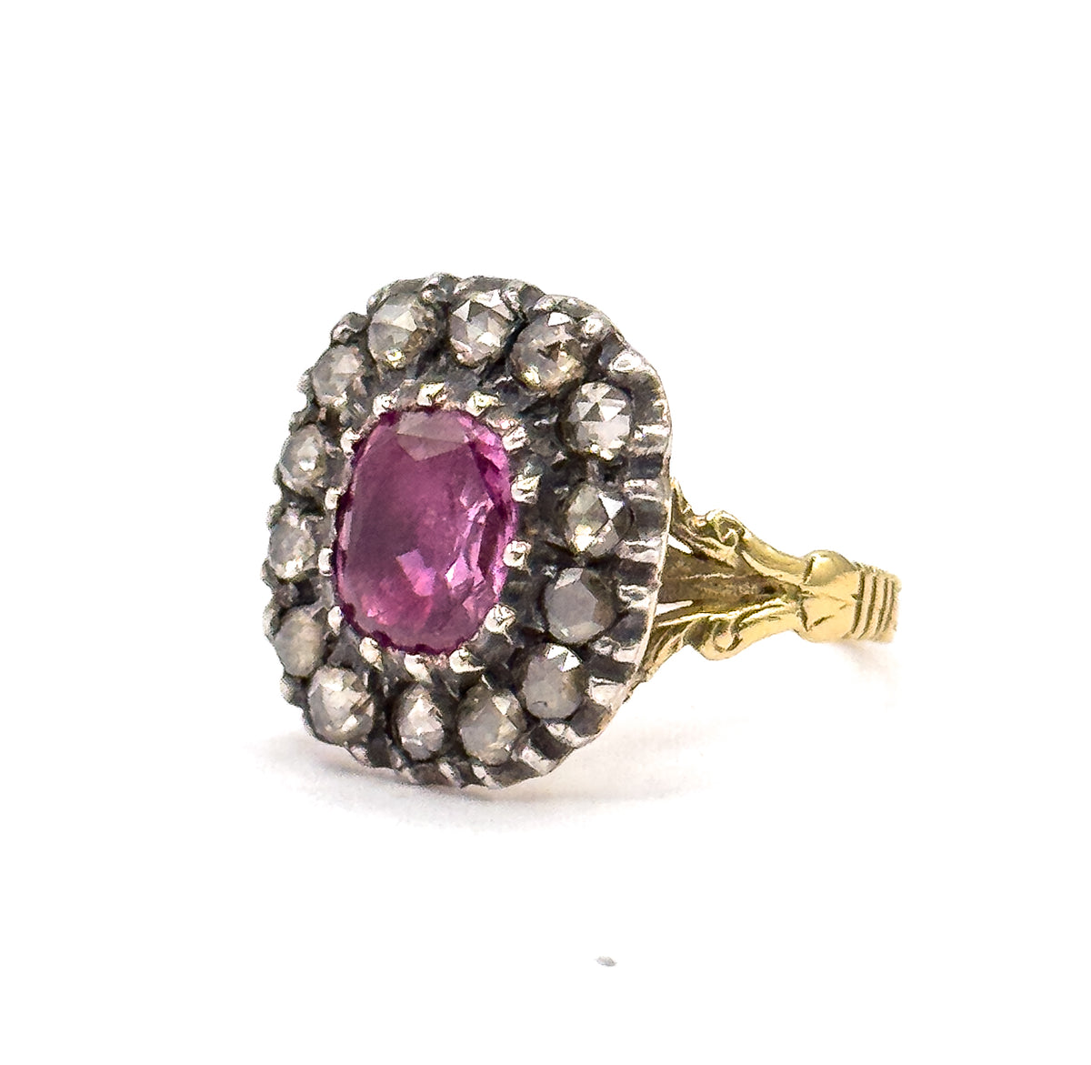 Turned three-quarters to the right, the full cushion-cut pink sapphire faces the viewer within its double row of silver-set diamonds, the foliate shoulders and grooved gold shank trailing behind.