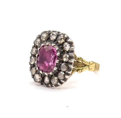 Turned three-quarters to the right, the full cushion-cut pink sapphire faces the viewer within its double row of silver-set diamonds, the foliate shoulders and grooved gold shank trailing behind.