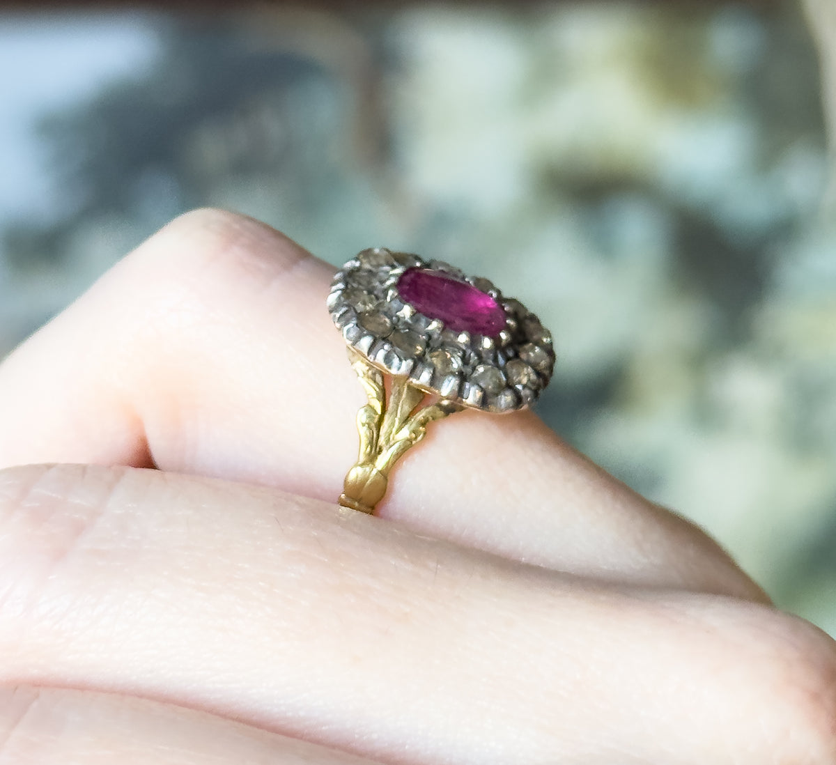 A closer three-quarter view on the finger, where the pink sapphire reads as a glowing oval against the pale silver-set diamond halo, the gold shoulders dropping away below.