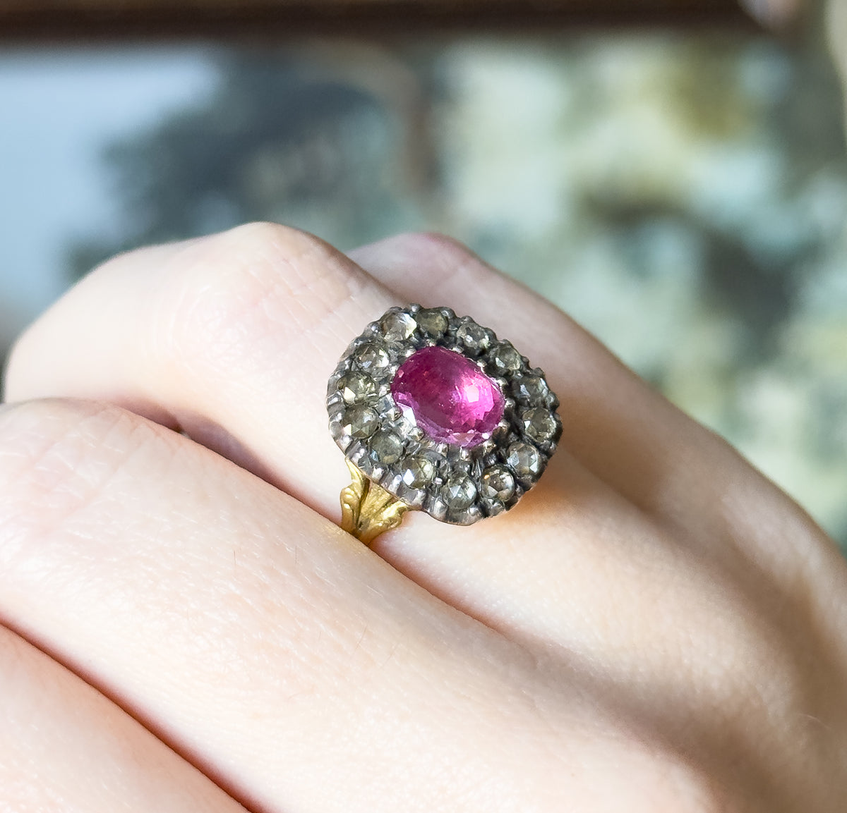 Seen from above on the finger, the full face shows the cushion-cut pink sapphire ringed by fourteen small diamonds in their silver setting, with one gold foliate shoulder just in view.