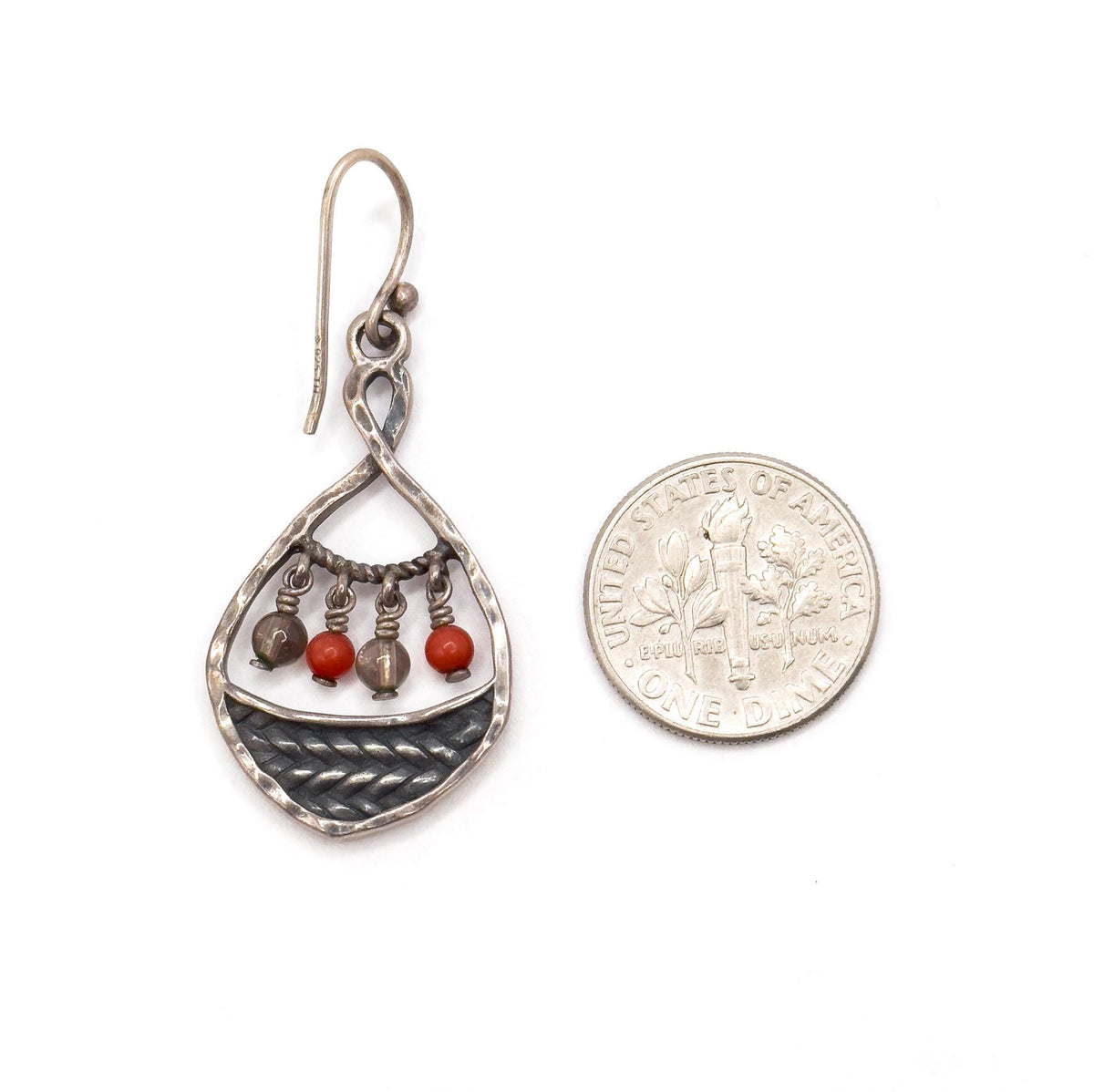 A single earring laid beside a dime for scale, the hammered teardrop frame about as wide as the coin and the whole drop roughly twice its height, braided dark panel and four beads facing up.