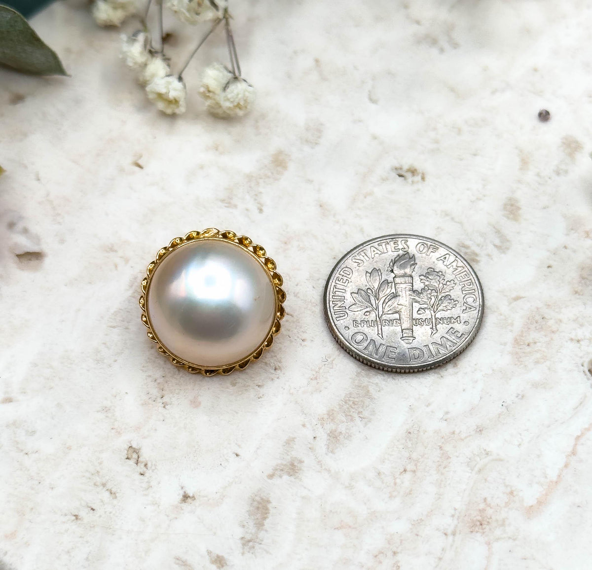 A single earring face-up beside a US dime for scale, the pearl and its rope twist bezel spanning a little more than half the coin's width, on the pale stone slab with dried flowers above.