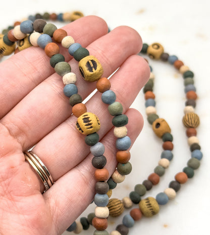 Strands draped across an open palm for scale — the small round beads are roughly a fingernail's width, the marked ochre beads a little larger — with the rest of the necklace curving onto white behind.
