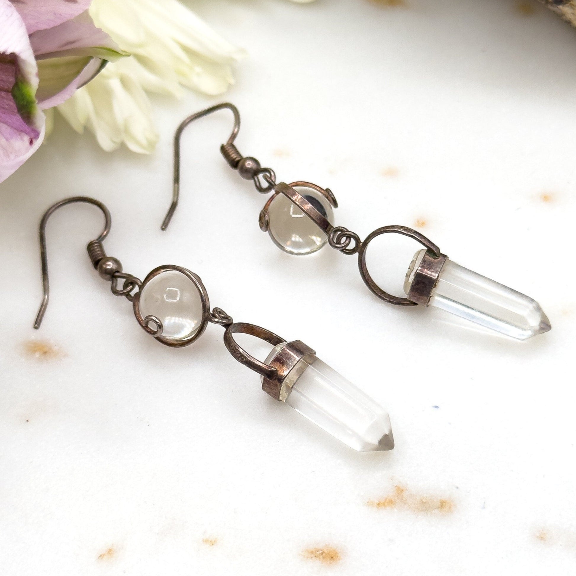 Pair of earrings with clear stones on a white surface with flowers and a crystal.