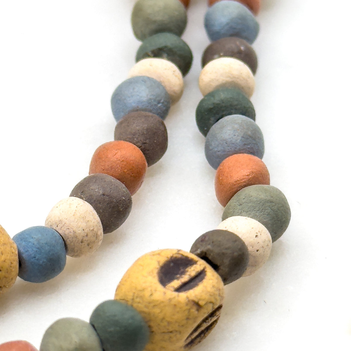Overhead close-up of two parallel rows running diagonally across white, showing the matte surface of each small round bead in blue, green, cream and rust, with a marked ochre bead at the lower edge.