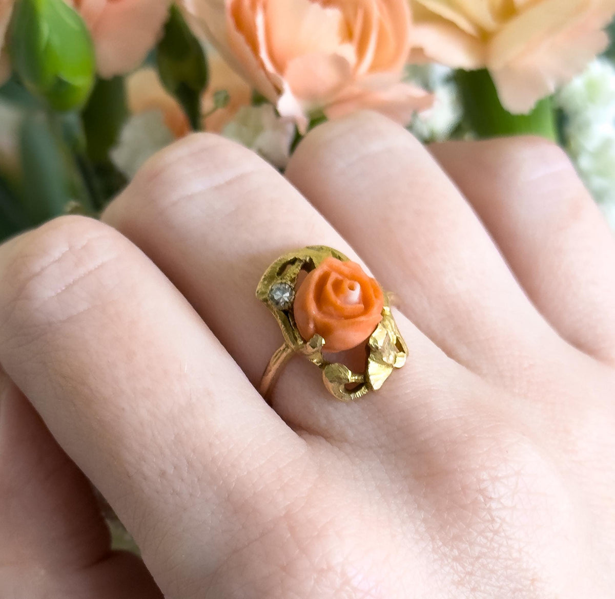 Almost the same view with slightly different angle, closer overhead on the finger, showing the leaf-like gold scrolls curling under the coral rose and the thin shank between the fingers.