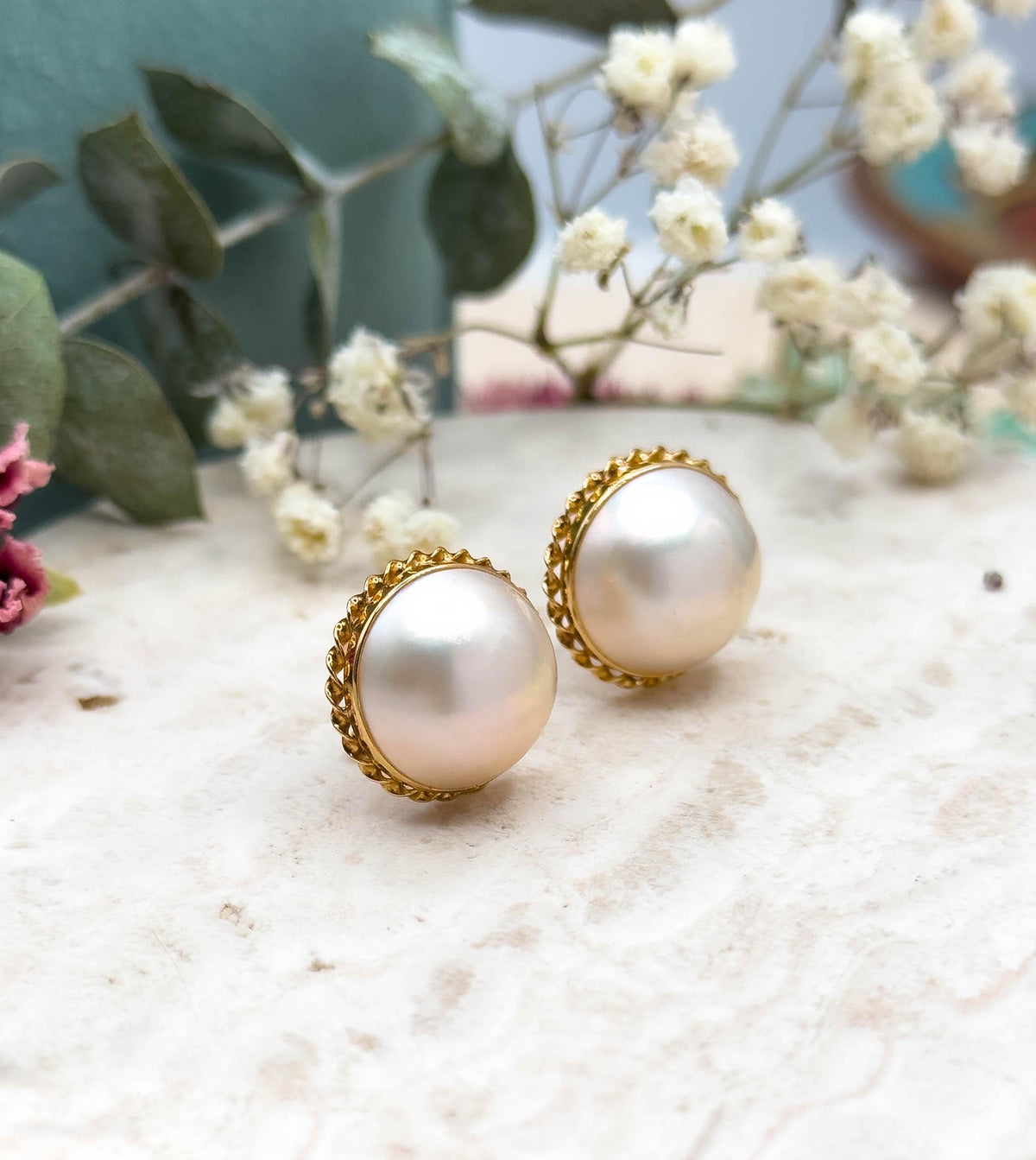 Three-quarter view with the pair standing upright on the stone slab, pink dried blooms at left, showing the depth of each pearl dome and the open gold gallery beneath the rope twist bezel.
