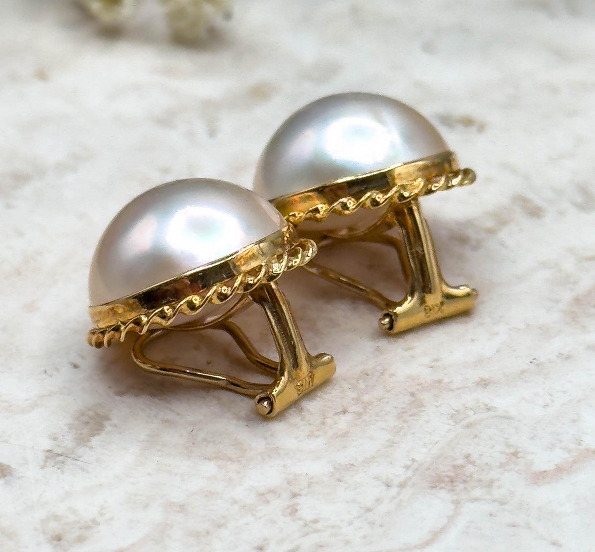 Low side view of the pair on the pale stone slab, the open clip levers standing up behind each bezel so the rope twist rim and the curve of the pearl domes are seen in profile.