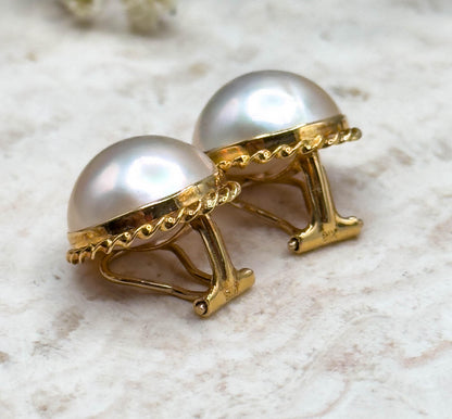 Low side view of the pair on the pale stone slab, the open clip levers standing up behind each bezel so the rope twist rim and the curve of the pearl domes are seen in profile.