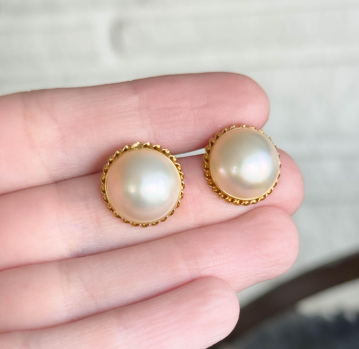 Both earrings face-up resting on a hand's fingertips for scale, each domed pearl in its rope twist yellow gold bezel spanning roughly the width of a fingertip.