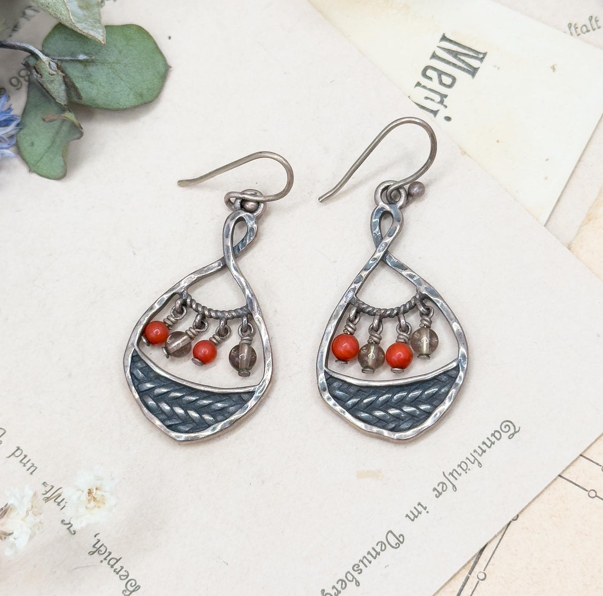 Both earrings upright side by side on the layered printed cards, showing the twisted silver loop each ear wire passes through and the fine rope-twist bar the coral and clear beads hang from.