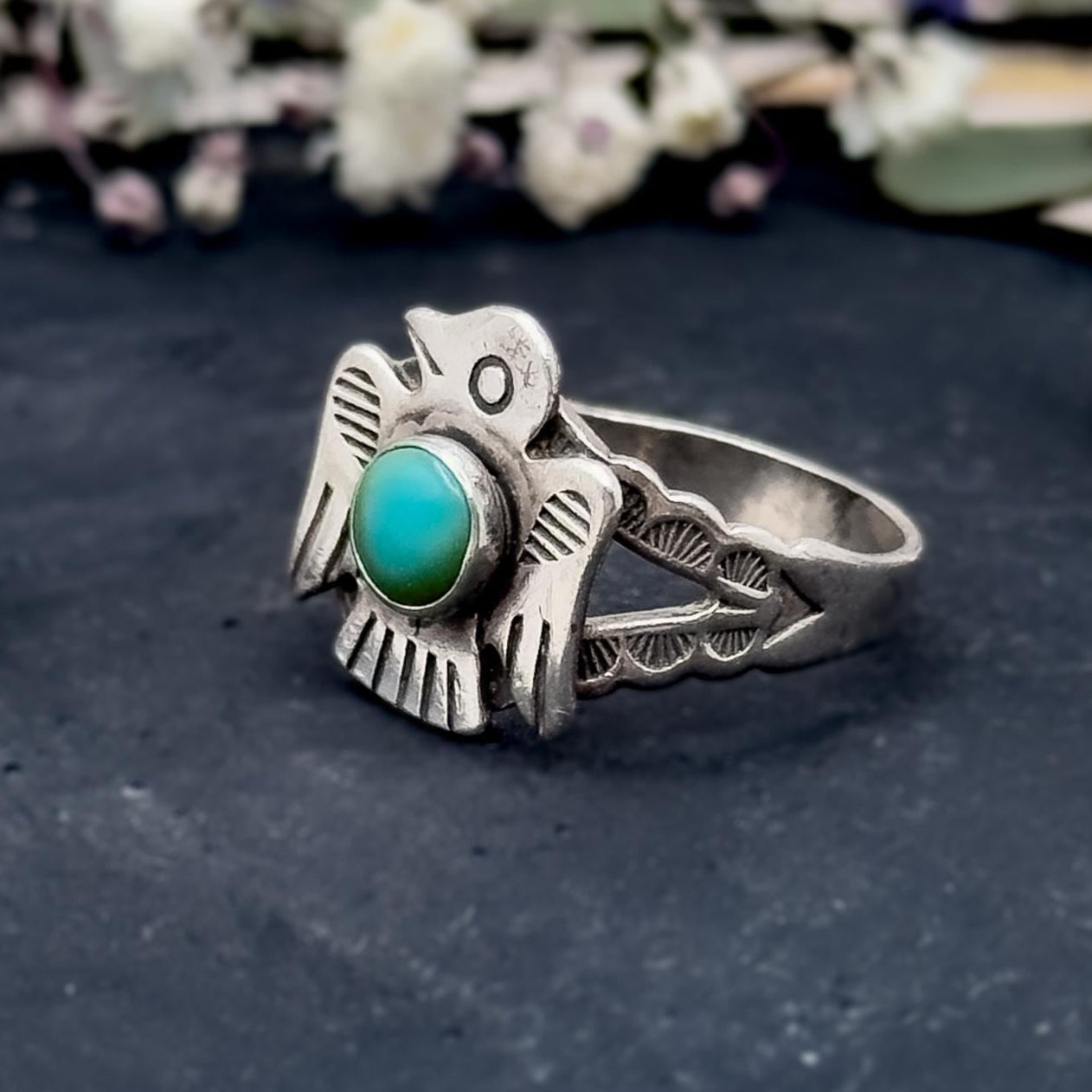 Silver ring with a turquoise stone on a dark surface with flowers in the background